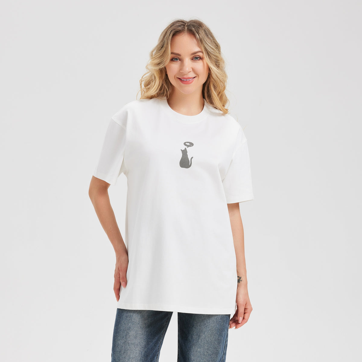 A woman modeling an oversized white t-shirt with a small black cat graphic on the chest, paired with blue jeans, against a light gray studio background.